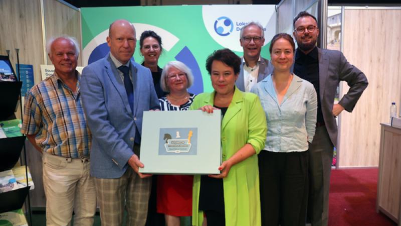 Philip van Veller en Sharon Dijksma houden een bord vast met het logo van het spel, om hen heen VNG-medewerkers die hebben bijgedragen aan het spel